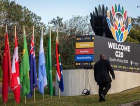 لافتة ترحيبية لقمة مجموعة العشرين (G20) في جنوب أفريقيا محاطة بأعلام الدول المشاركة، مع رجل يسير باتجاه موقع الحدث. - بلومبرغ