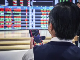 شاشة هاتف ذكي تعرض تحركات الأسهم في يد شخص يقف أمام شاشة تعرض أسعار الأسهم - بلومبرغ