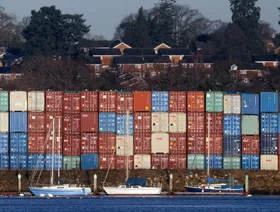 مجموعة حاويات مكدسة في انتظار عمليات الشحن والتفريغ وسفن صغيرة ترسو أمامها - المصدر: بلومبرغ