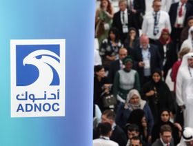 أشخاص يرتدون ملابس متنوعة بعضهم يرتدي الزي الوطني الإماراتي بجوار لافتة تحمل شعار شركة بترول أبوظبي الوطنية (أدنوك) - بلومبرغ