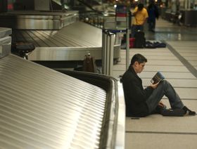 رجل يقرأ كتاباً وهو جالس مسنداً ظهره إلى ناقل الحقائب في قاعة الوصول بأحد المطارات - المصدر: غيتي إيمجز
