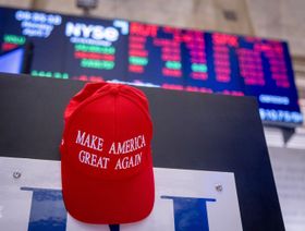 قبعة حمراء تحمل شعار "اجعلوا أميركا عظيمة مجدداً" (Make America Great Again) المرتبط بالرئيس الأميركي دونالد ترمب، معلقة داخل قاعة التداول في بورصة نيويورك، وتظهر في الخلفية شاشات تعرض بيانات الأسواق المالية والتي يغلب عليها اللون الأحمر، ما قد يشير إلى تراجع في الأسهم. الصورة تبرز تداخلاً رمزياً بين السياسة والأسواق المالية الأميركية. - بلومبرغ
