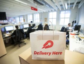 كيس ورقي يحمل شعار شركة "دليفري هيرو" (Delivery Hero)، المتخصصة في خدمات توصيل الطعام، موضوع على منضدة داخل مكتب. ويظهر في الخلفية عدد من الموظفين مشغولين بأداء مهامهم على أجهزة الكمبيوتر. - المصدر: بلومبرغ