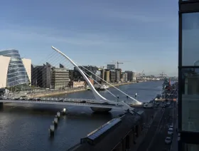 جسر في العاصمة الأيرلندية دبلن يمتد فوق مياه نهر هادئ تحيط به مبانٍ حديثة ذات واجهات زجاجية عاكسة لأشعة الشمس - بلومبرغ