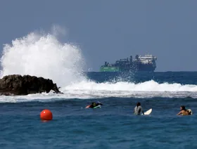 أشخاص يمارسون رياضة ركوب الأمواج، ويظهر في عمق الصورة سفينة تشارك في مد خط أنابيب لنقل الغاز الطبيعي في البحر الأبيض المتوسط بالقرب من شاطئ دور، شمال إسرائيل، 28 أغسطس 2018. - رويترز