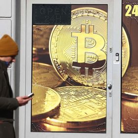 شخص يطالع هاتفه المتحرك يعبر من أمام متجر تزينه صورة لقطع رمزية للعملة المشفرة "بتكوين" - المصدر: بلومبرغ