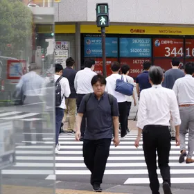 مشاة يعبرون الطريق في منطقة تجارية، ويظهر في الخلفية لوحات إلكترونية تعرض حركة مؤشر "نيكاي 225" والأسهم، في واجهة شركة للأوراق المالية، طوكيو، اليابان - المصدر: بلومبرغ