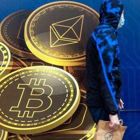 شخص يرتدي كمامة يعبر من أمام لوحة إعلانية تحمل شعارات عملتي "بتكوين" و"إيثريوم" المشفرتين - المصدر: بلومبرغ