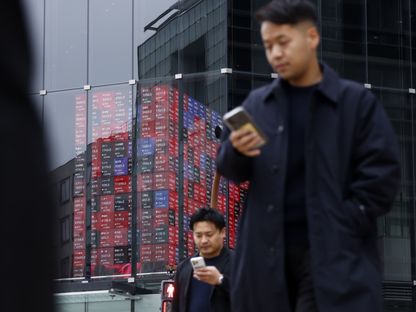 مارة يطالعون هواتفهم الذكية خلال سيرهم أمام مبنى كابوتو وان، وتبدو داخله لوحة إلكترونية تعرض أسعار أسهم. طوكيو، اليابان - المصدر: بلومبرغ
