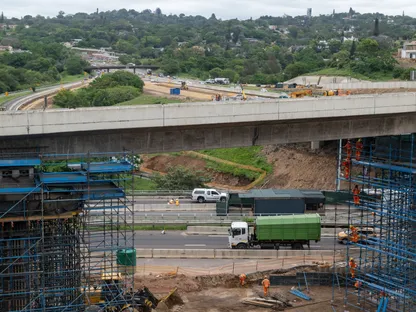 عمال يرتدون ملابس برتقالية يعملون على سقالات معدنية زرقاء في منطقة واسعة تشهد أعمال بناء ضخمة للبنية التحتية - بلومبرغ