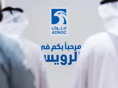 شعار \"أدنوك\" على جدار ويظهر في المقدمة رجلان يرتديان الزي الإماراتي التقليدي وشخص ثالث من جهة اليسار يرتدي زي عمل أزرق في مصفاة تابعة للشركة في الرويس. - بلومبرغ