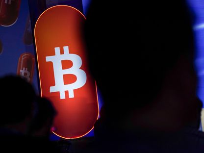 شعار بتكوين على خلفية برتقالية متوهجة ضمن لوحة مضيئة في معرض للعملات المشفرة - بلومبرغ