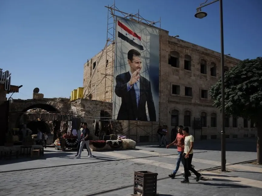 أشخاص يمرون أمام صورة للرئيس السوري بشار الأسد - المصدر: بلومبرغ